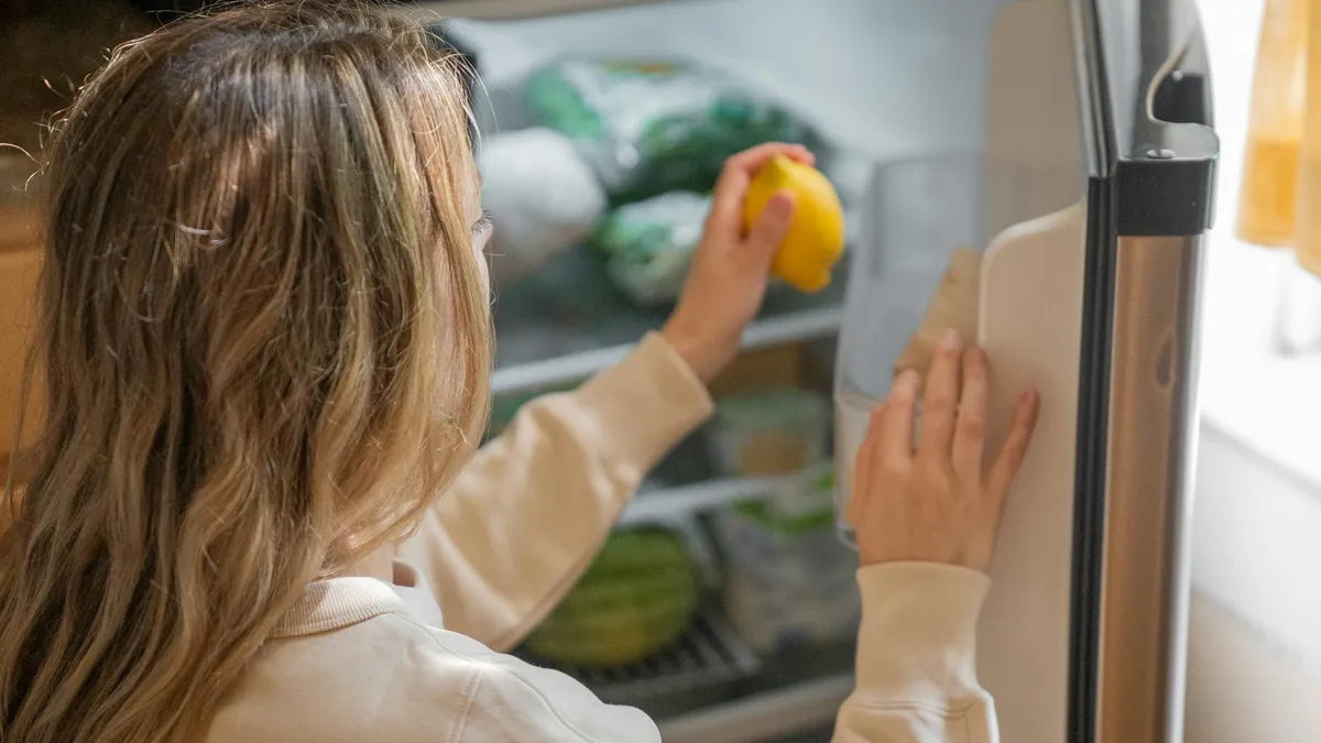 Person reaching for a lemon inside an open refrigerator stocked with fresh vegetables and herbs